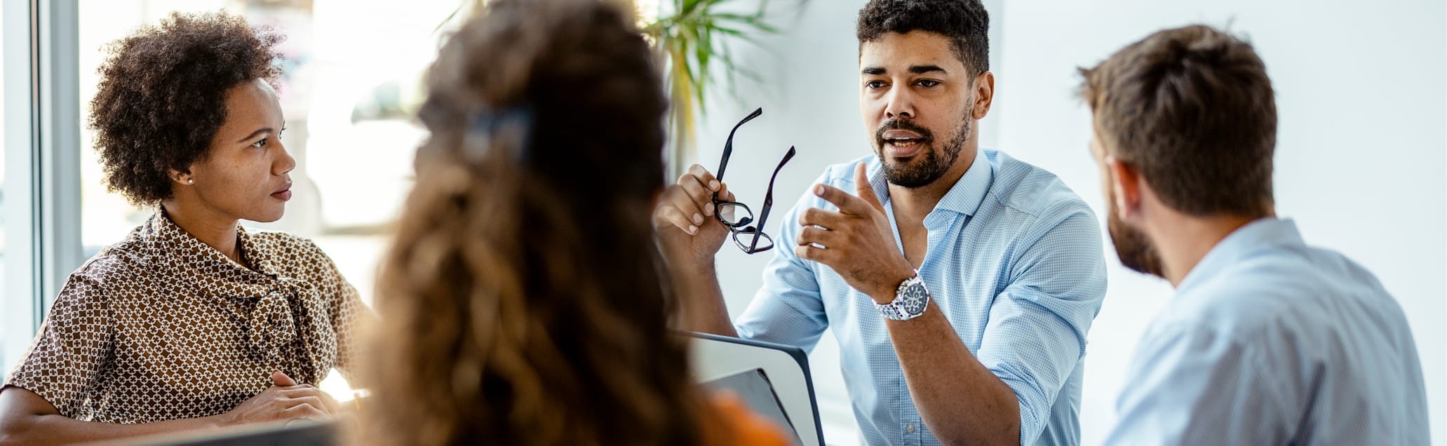 A man leading a meeting with three other people while holding his glasses